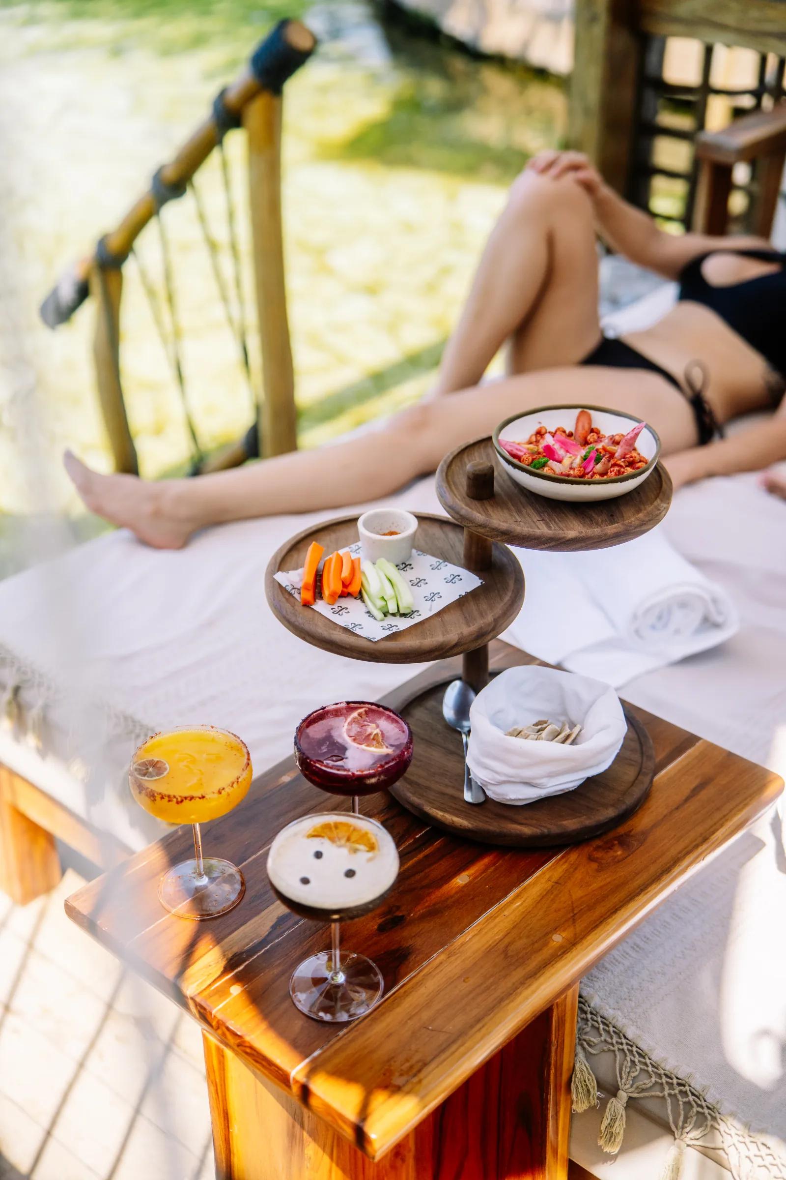 Cocktails and snacks sit on a table beside a person lounging on a sunbed at Atta / Tuuch.