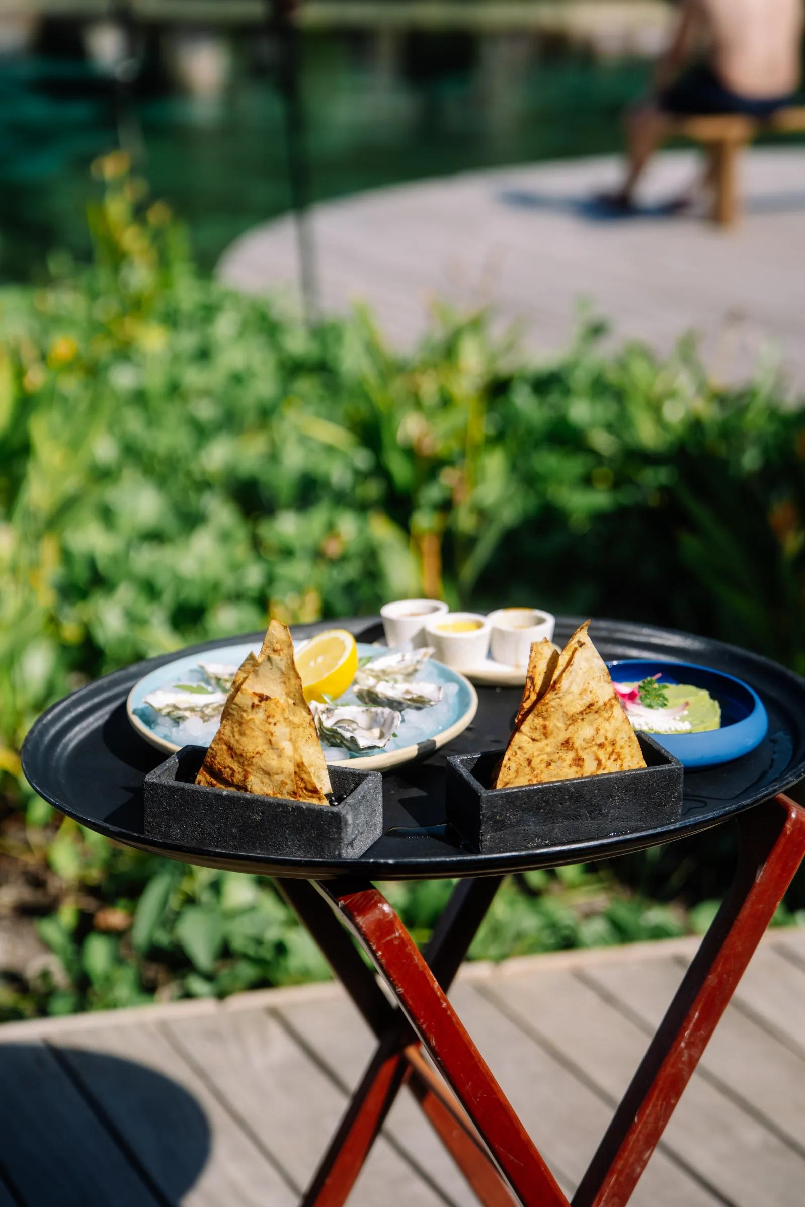 Fresh oysters and appetizers sit on a tray on Atta's sunny outdoor deck near lush foliage.