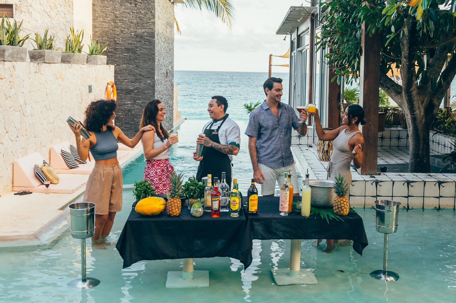 Five people gather around a bar table in shallow turquoise water at Mi Amor, with tropical plants and ocean views behind