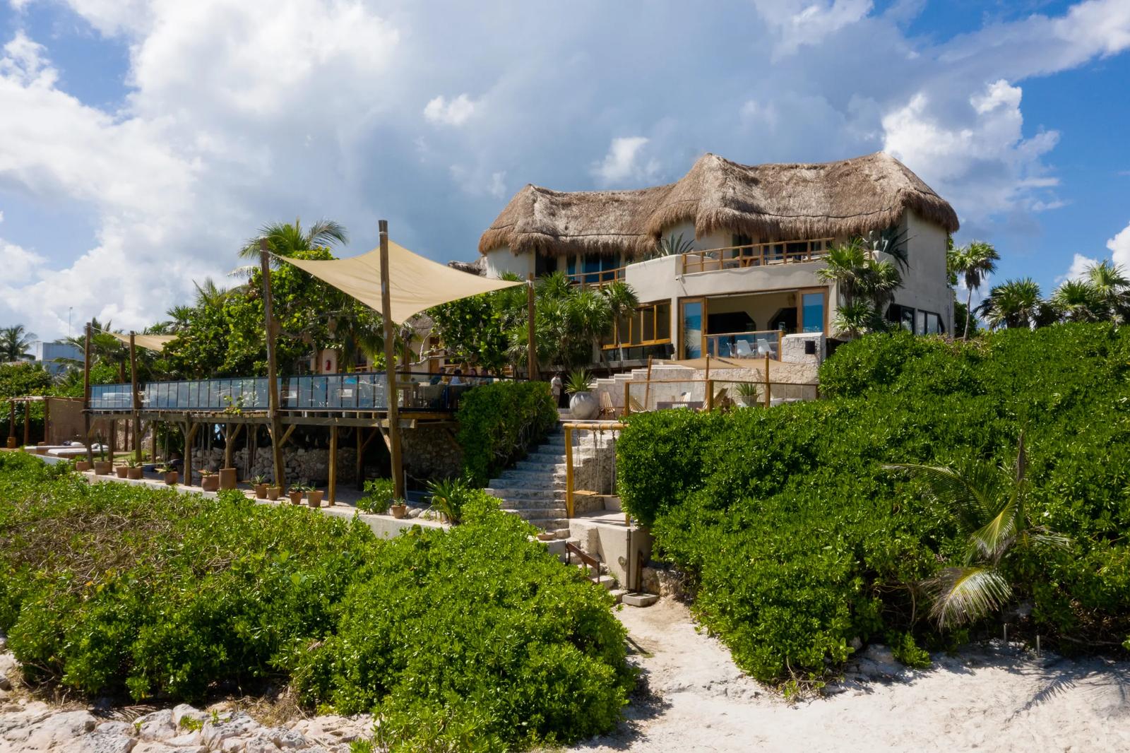 Mezzanine beach club with thatched-roof structures, wooden decks, and canvas shade sails surrounded by tropical vegetation