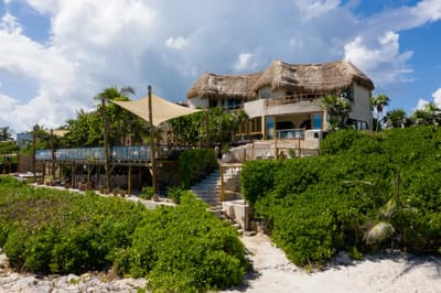 Mezzanine beach club with thatched-roof structures, wooden decks, and canvas shade sails surrounded by tropical vegetation