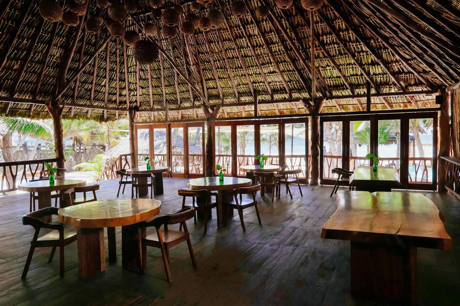 Tulumania’s open-air dining room overlooks a palm-lined beach, with rustic tables beneath a thatched roof.