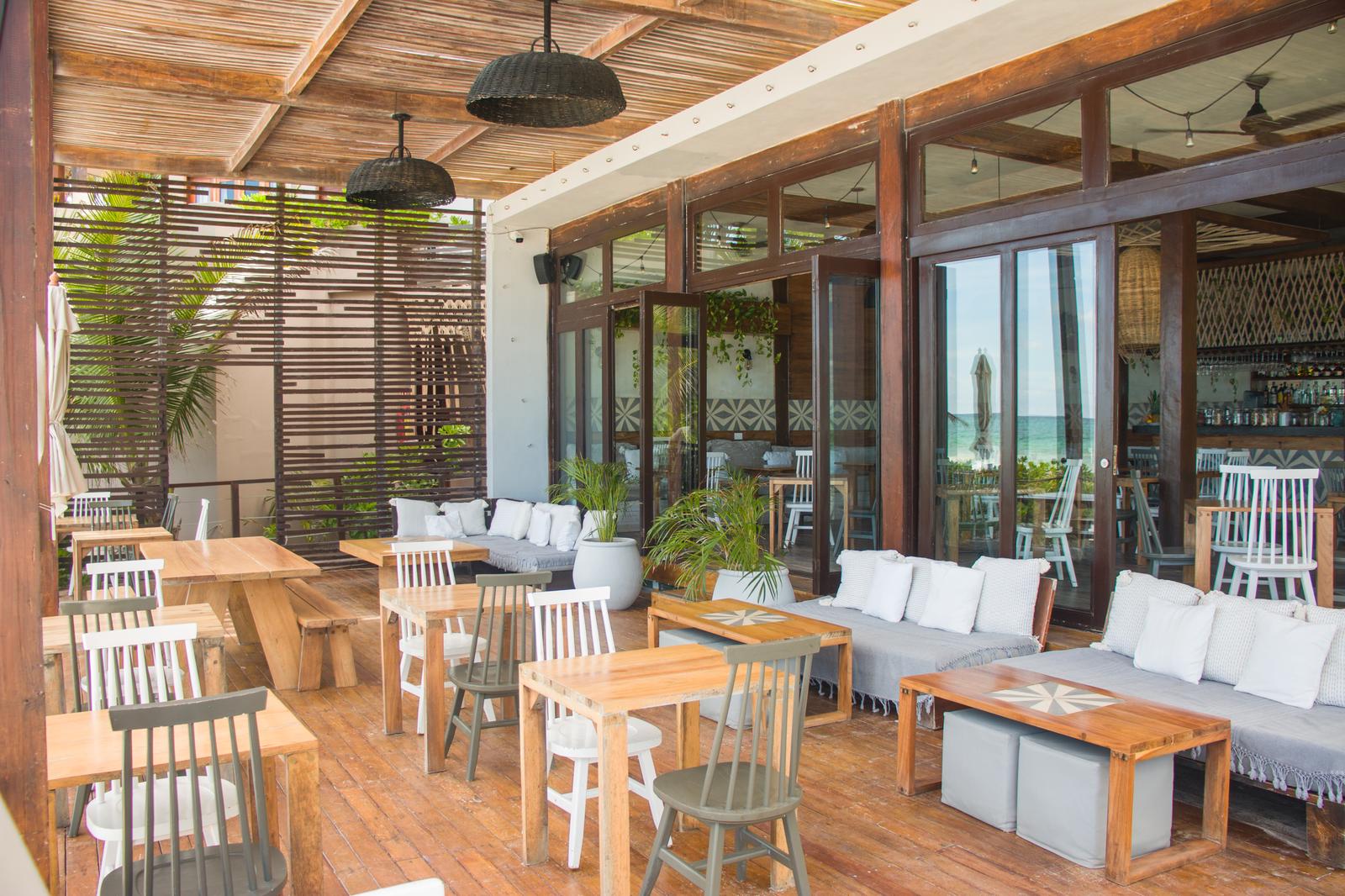La Zebra Mediterranean terrace with wooden tables, white chairs, lounge seating, and ocean views through glass doors.