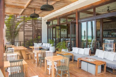La Zebra Mediterranean terrace with wooden tables, white chairs, lounge seating, and ocean views through glass doors.