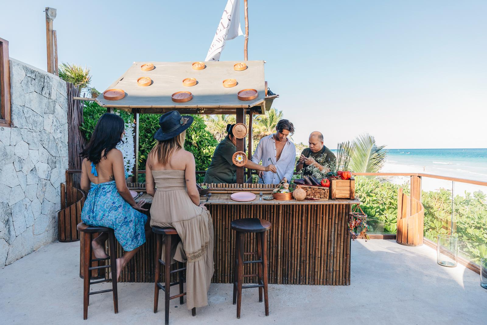 Guests enjoy drinks and food at Mezzanine's beachfront bar with ocean views and tropical vegetation in the background.