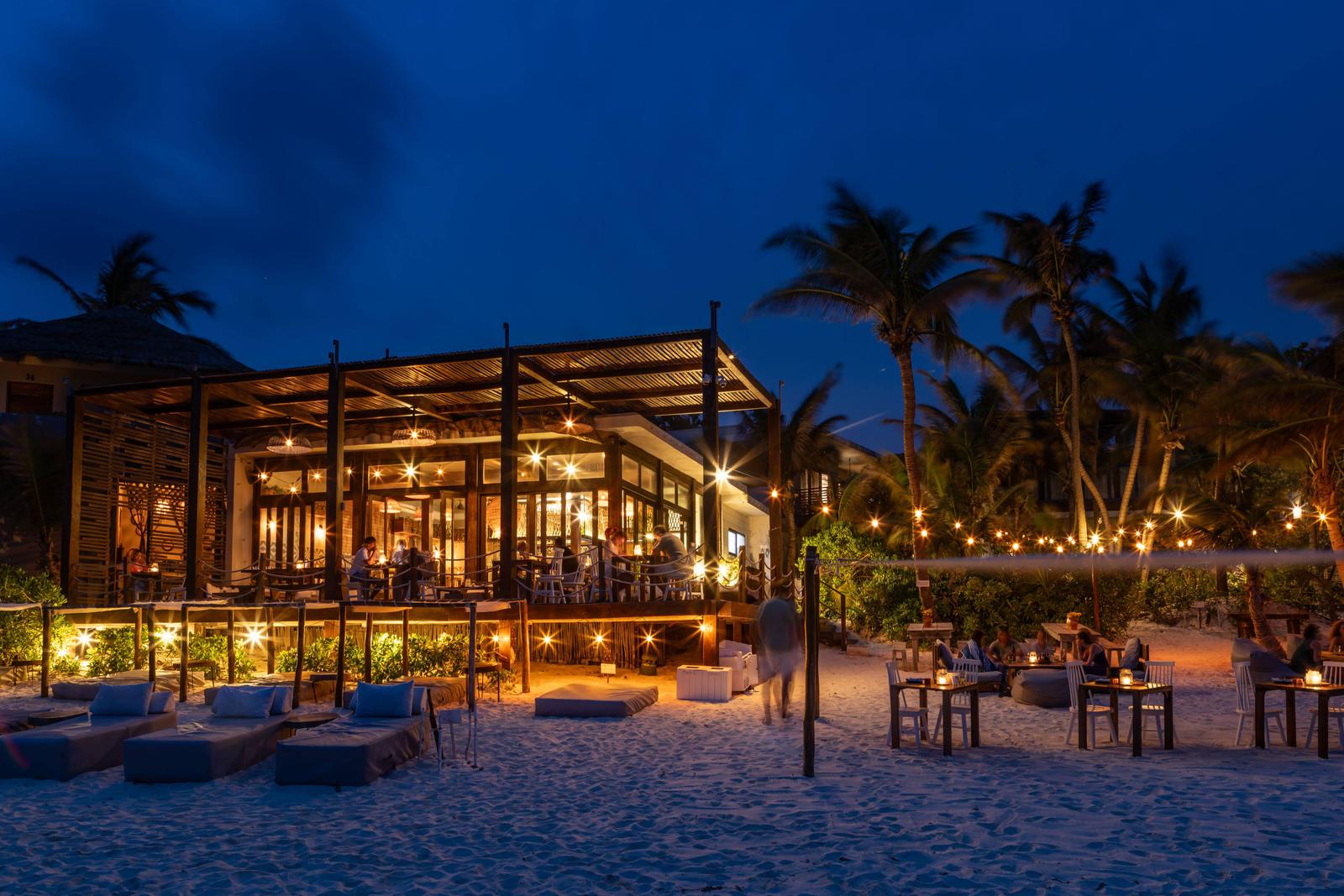 La Zebra Mediterranean beach club at dusk with illuminated dining pavilion, lounge seating, and palm trees along the sand.
