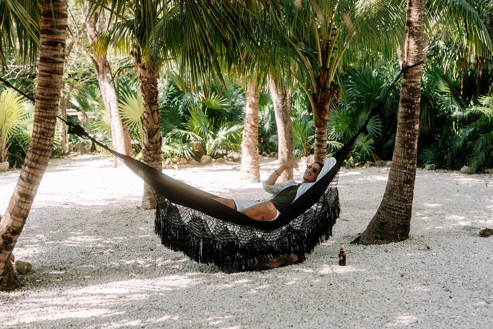 Person relaxing in a fringed hammock strung between palm trees on sandy ground surrounded by tropical vegetation.