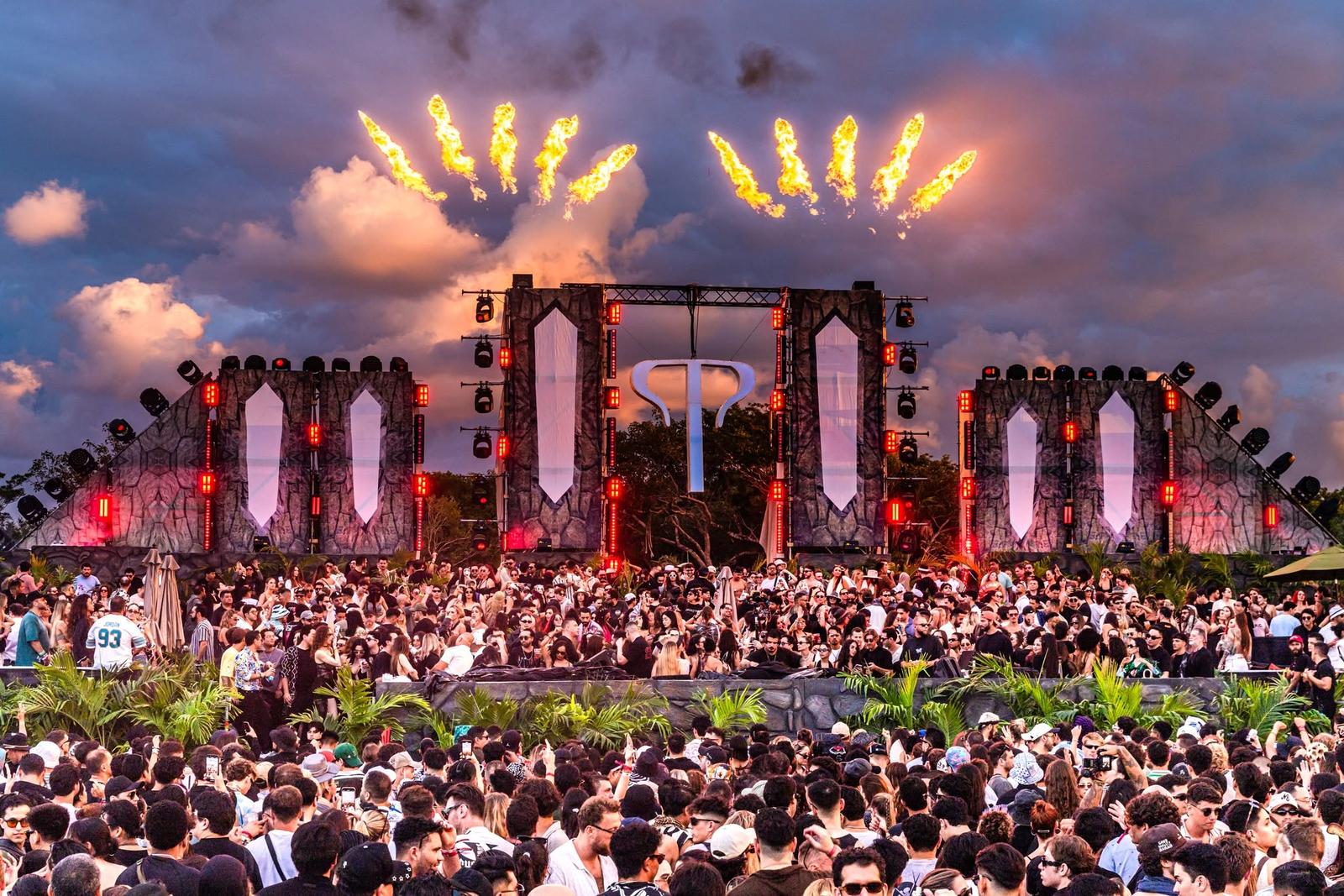 A large crowd gathers before a lit outdoor concert stage as fireworks burst overhead at dusk.
