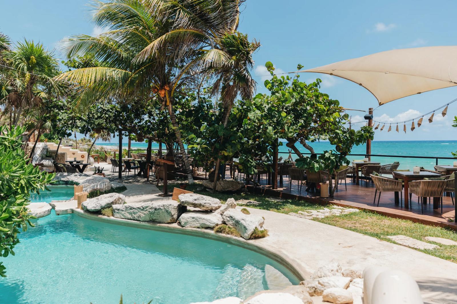Mezzanine's beachfront terrace with turquoise pool, palm trees, dining tables, and Caribbean Sea views under clear sky.