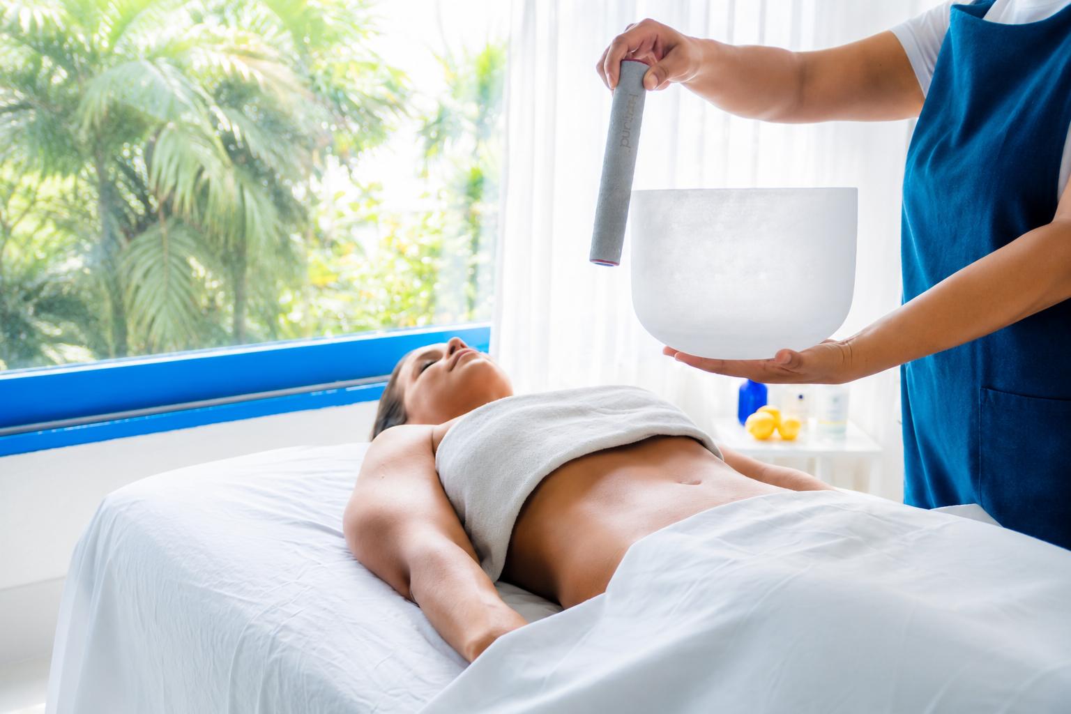 Therapist performing sound bath healing with singing bowl on client lying on massage table in bright room with ocean view