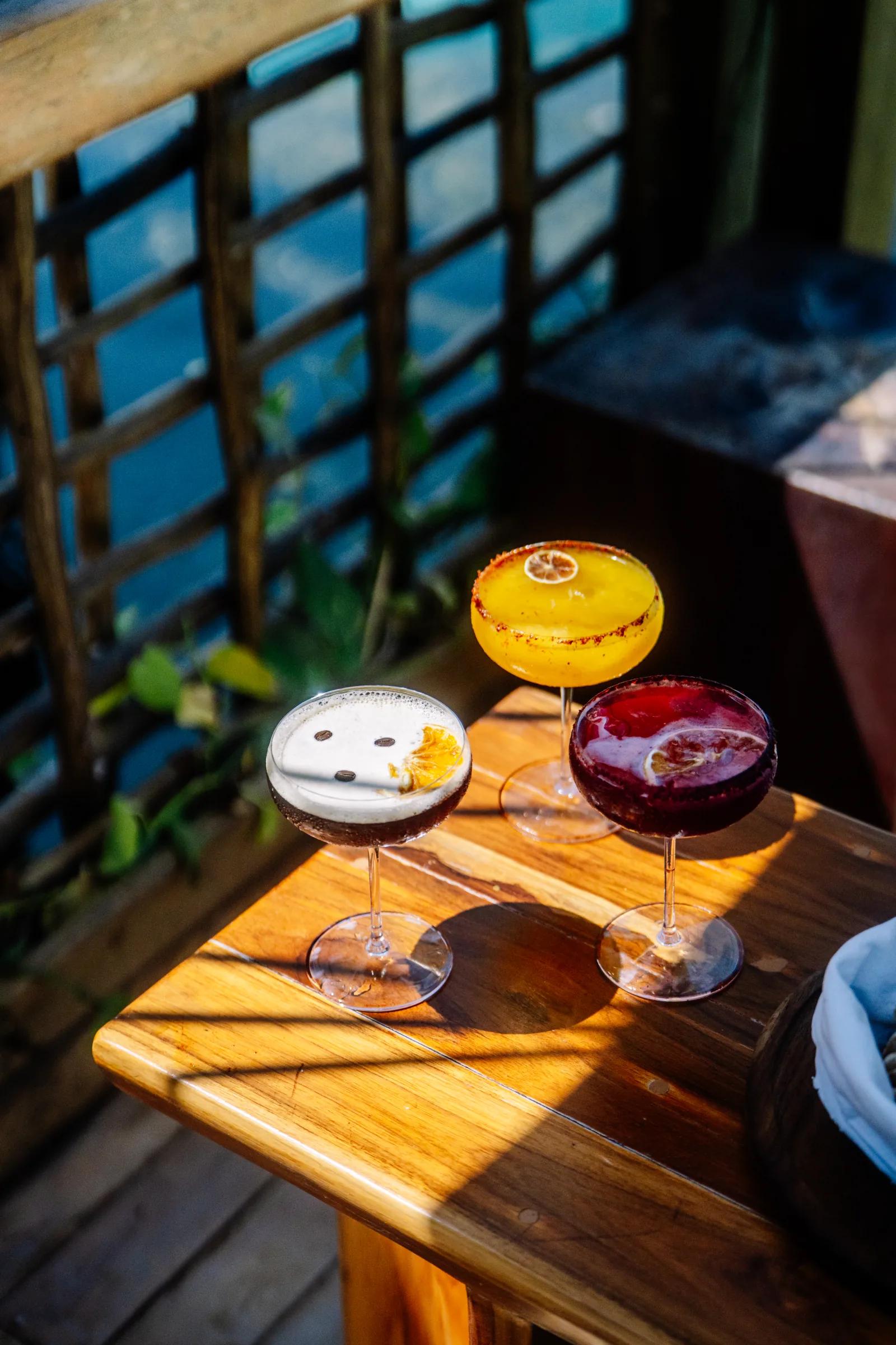 Three colorful cocktails rest on a sunlit wooden table on Atta's outdoor deck.
