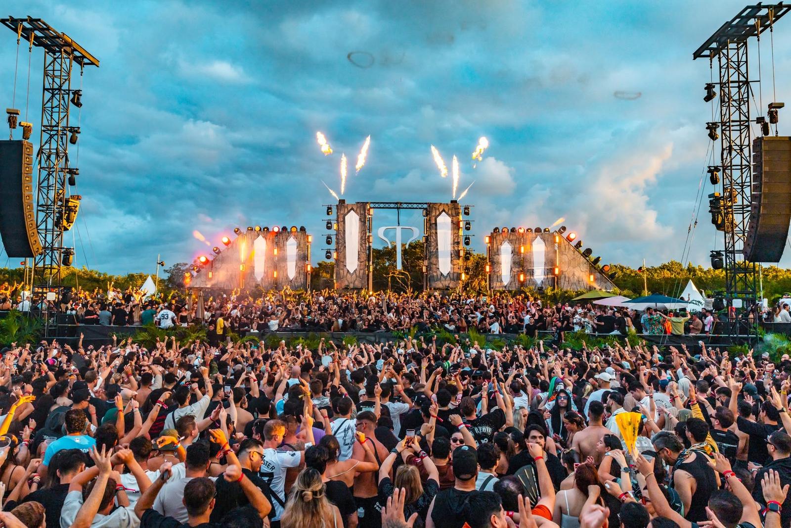 Outdoor music festival stage with fireworks and a large cheering crowd under a cloudy blue sky.
