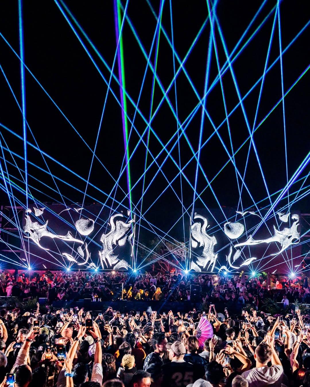 Nighttime crowd watches laser beams and projected figures above a stage, creating an energetic atmosphere.