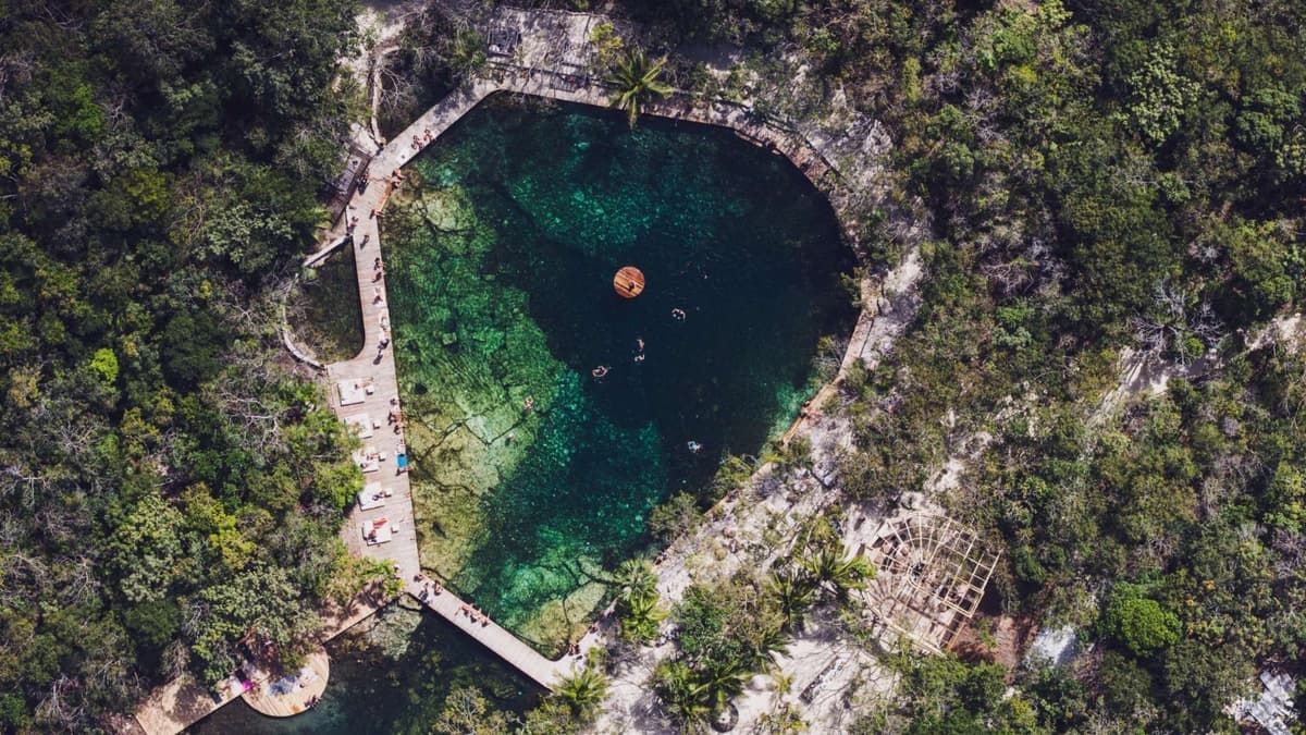 Aerial view of a turquoise cenote surrounded by jungle, stone paths, loungers, and thatched structures in Tulum.