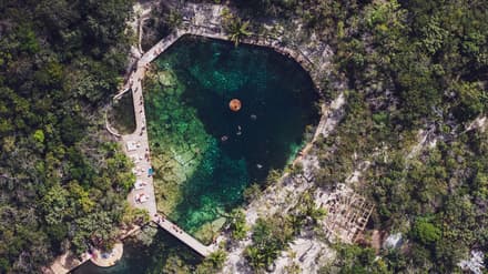 Aerial view of a turquoise cenote surrounded by jungle, stone paths, loungers, and thatched structures in Tulum.