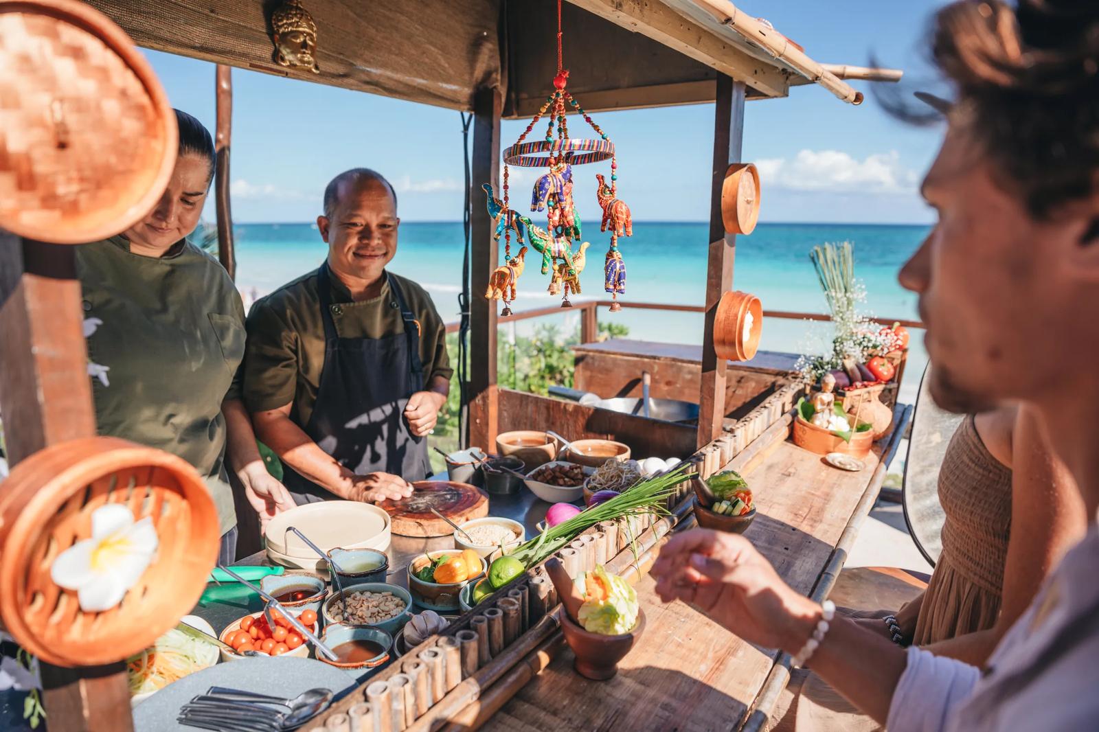 Chefs prepare food at Mezzanine's beachfront dining setup with turquoise ocean and sky visible beyond the wooden terrace.
