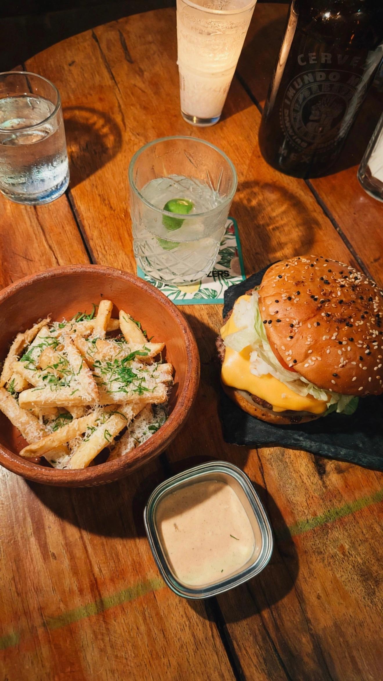 Burger, fries, dipping sauce, cocktails, and a beer on a wooden restaurant table create a casual dining mood.