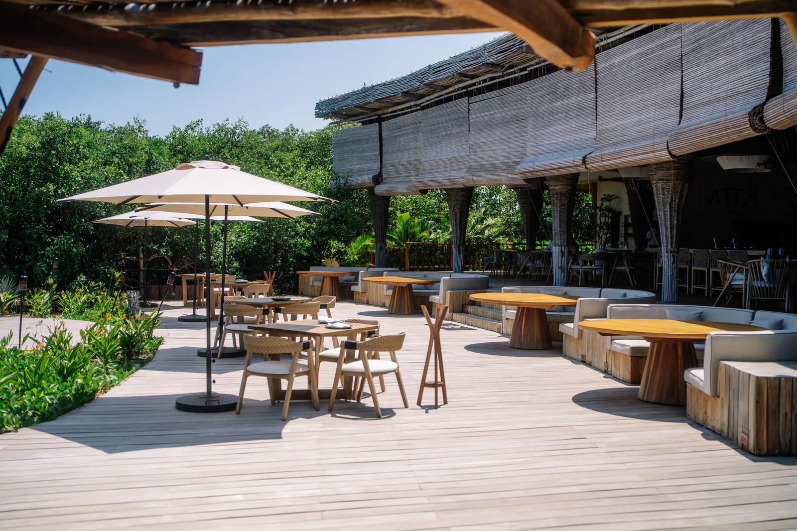 Tables, chairs, and umbrellas line the sunny wooden deck of Atta / Tuuch beside dense tropical greenery
