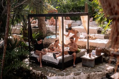 Guests relax on shaded daybed lounges surrounded by tropical vegetation at a beachside venue.