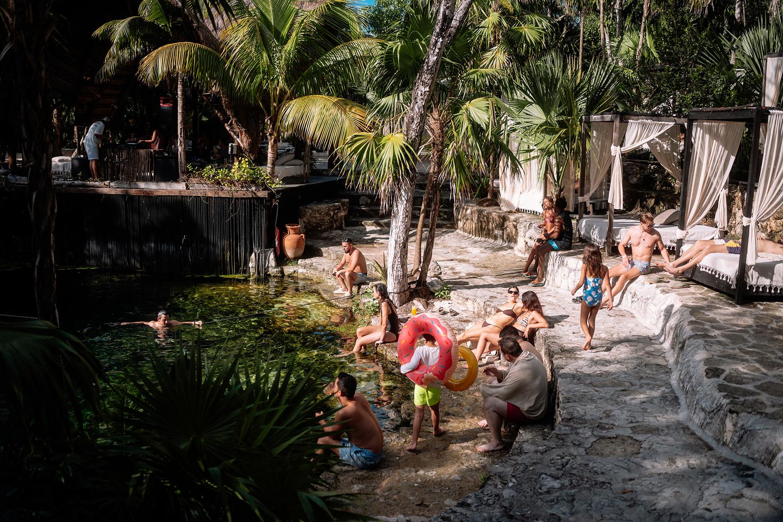 Guests swimming and relaxing around a natural cenote pool surrounded by palm trees and white cabanas.