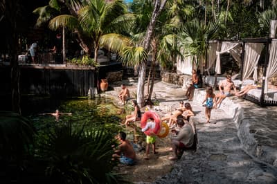 Guests swimming and relaxing around a natural cenote pool surrounded by palm trees and white cabanas.
