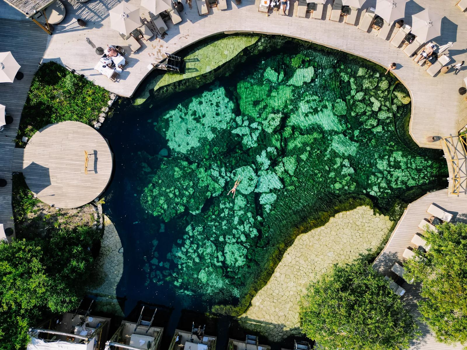 An aerial view shows a swimmer floating in the clear green cenote pool surrounded by wooden decks at Atta.