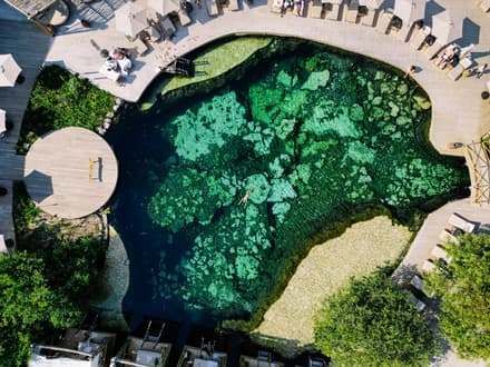 An aerial view shows a swimmer floating in the clear green cenote pool surrounded by wooden decks at Atta.