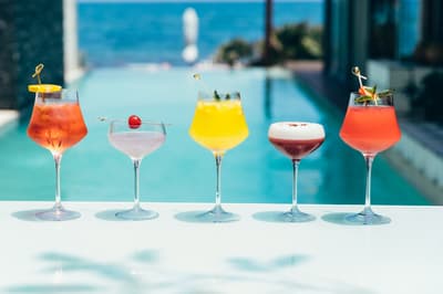 Five colorful cocktails lined up on a white counter with Mi Amor's pool and ocean view in the background.