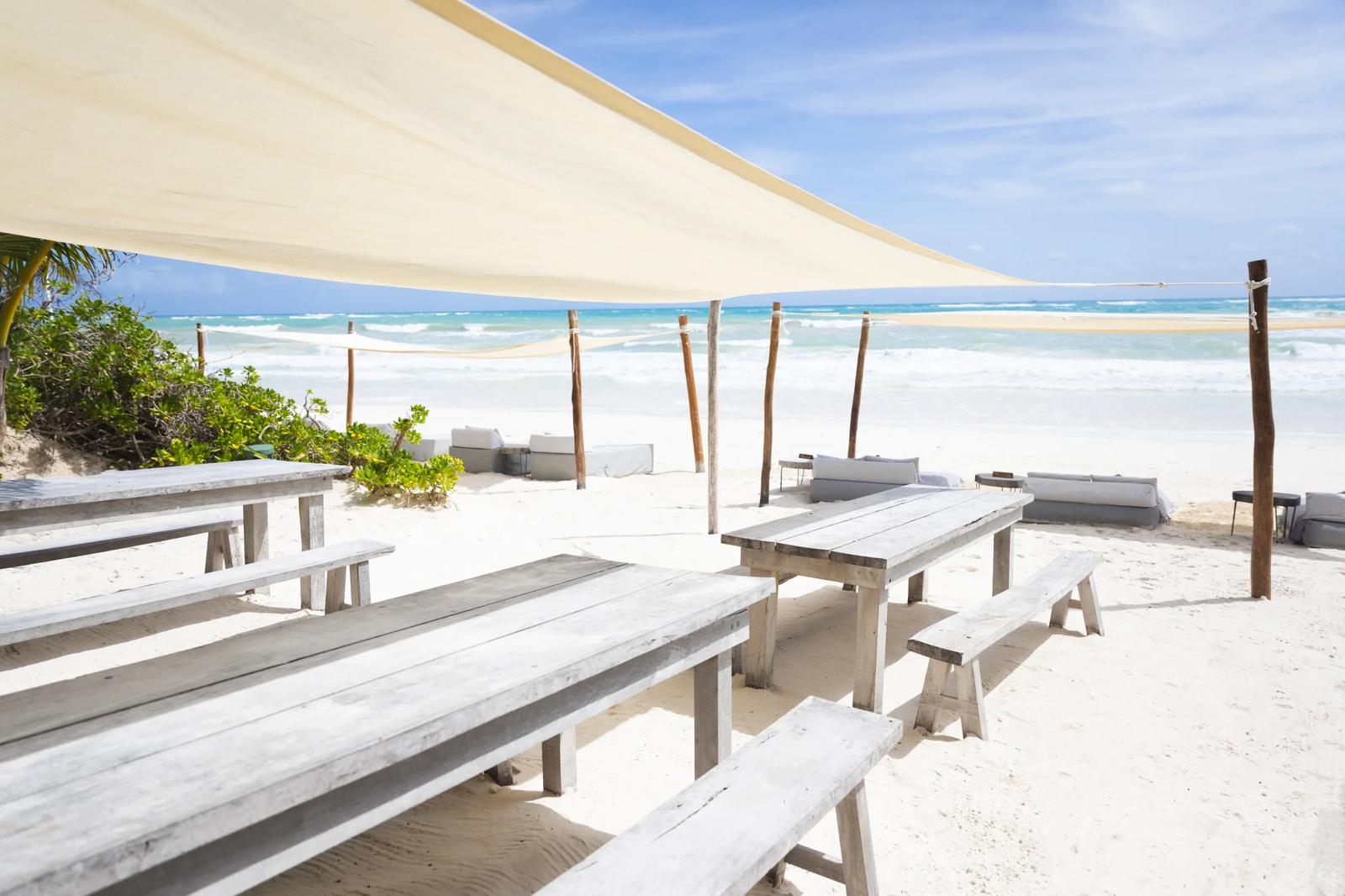 Wooden picnic tables sit on the white sand beach under shade sails at La Zebra Mediterranean, facing the ocean.