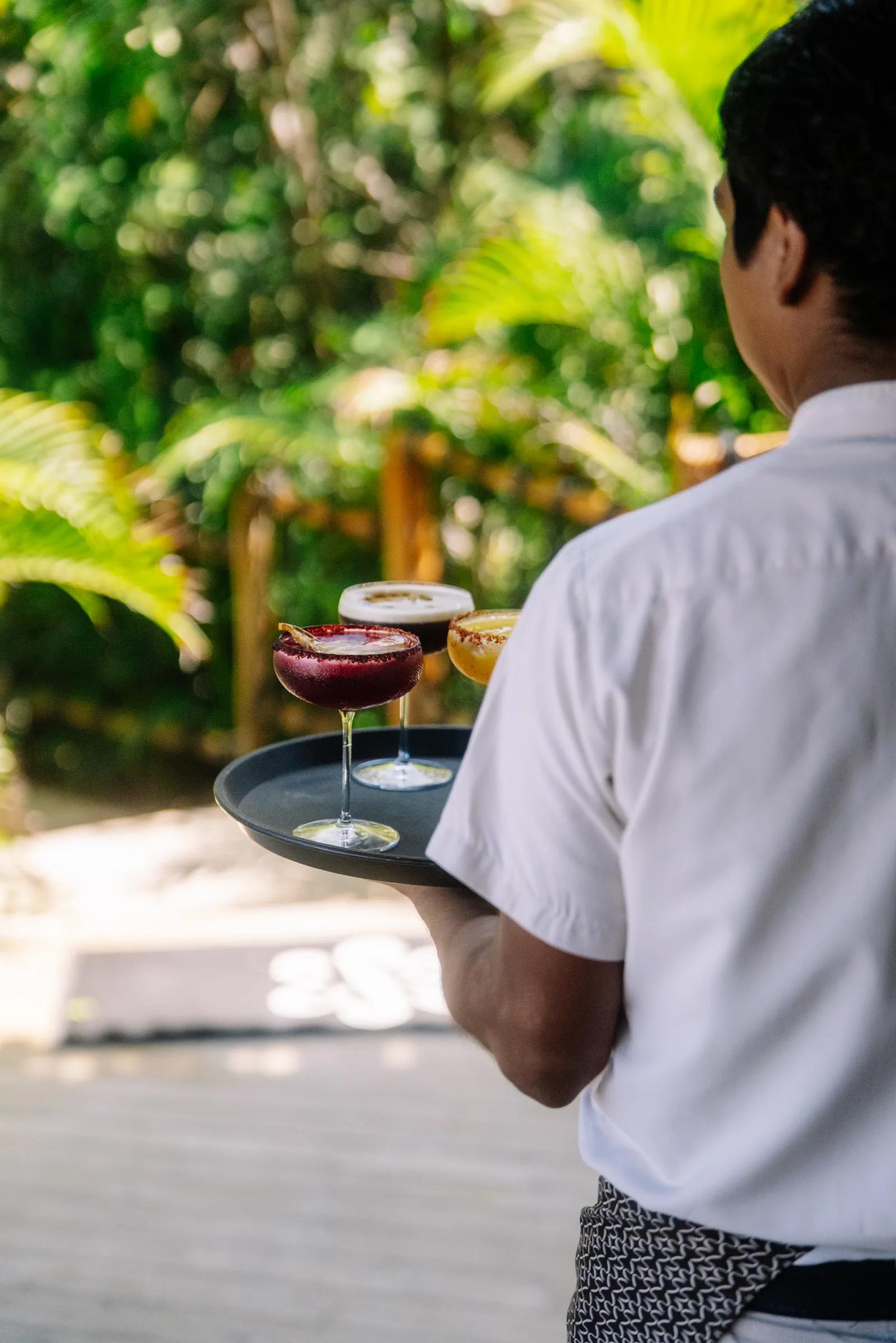 A waiter carries a tray of colorful cocktails outdoors at Atta against a backdrop of tropical greenery.