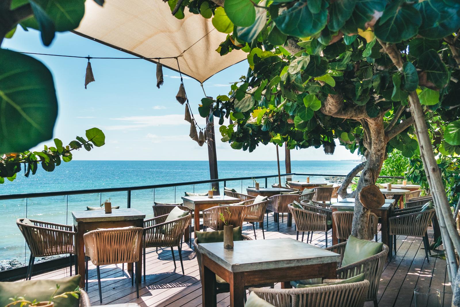 Mezzanine's beachfront terrace with wooden tables and woven chairs shaded by a canvas canopy and leafy trees, overlooking