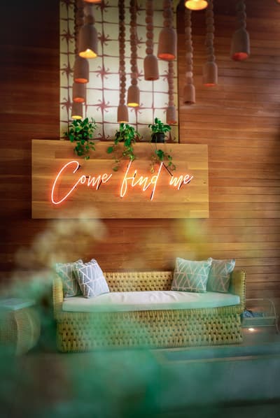 A woven sofa sits beneath a neon sign reading Come Find Me in a wood-paneled lounge area at Mi Amor.