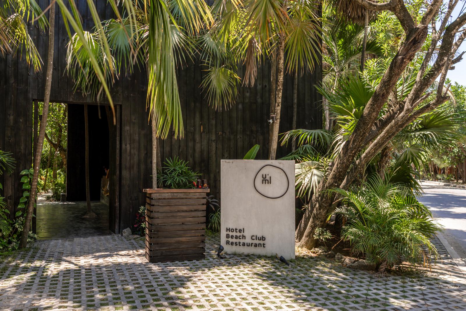 Umi Beachclub entrance sign beside a dark wooden building, surrounded by palms in a sunny tropical setting.