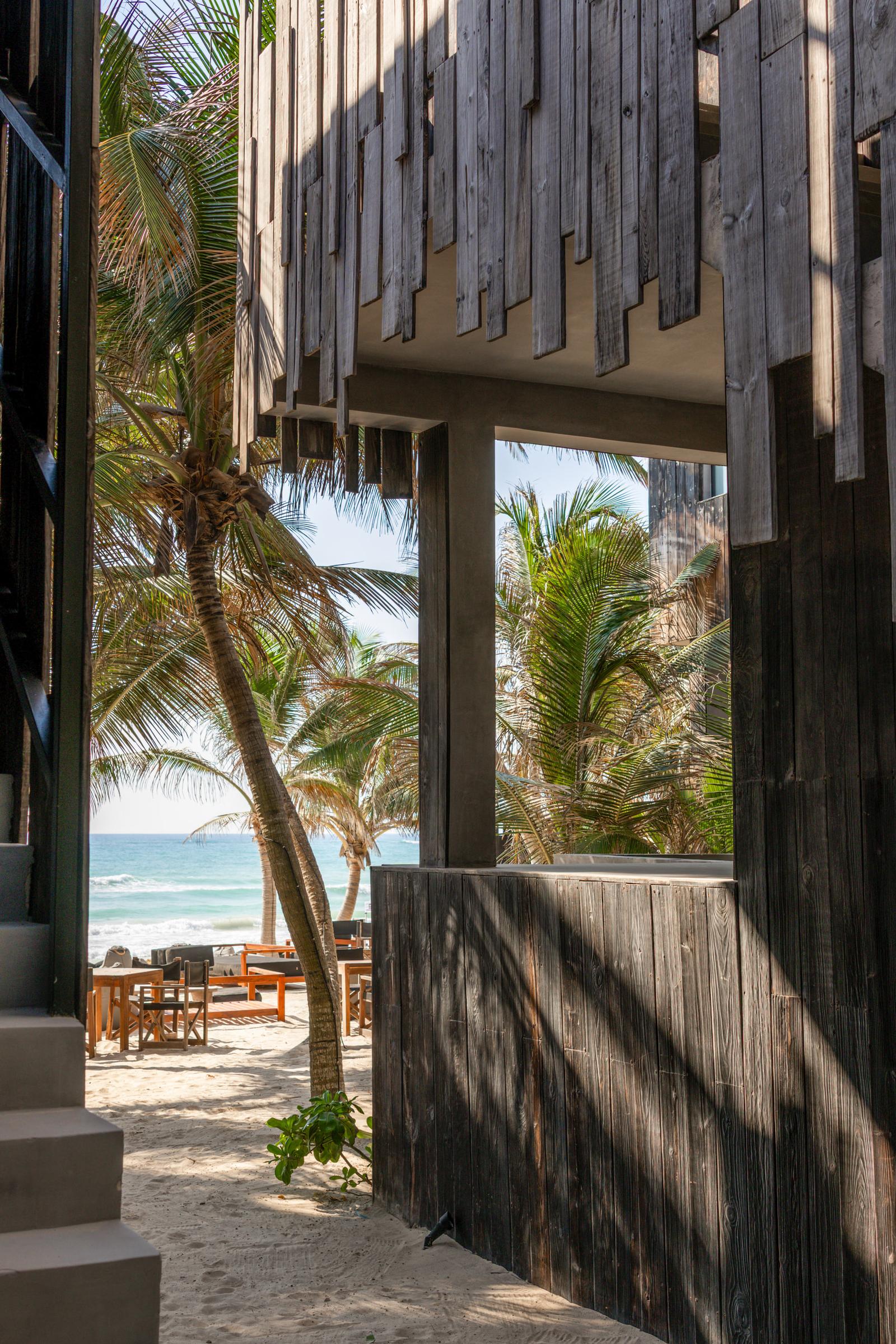 Wooden beach club entrance opens to palm trees, sandy seating, and turquoise ocean in bright sunlight.