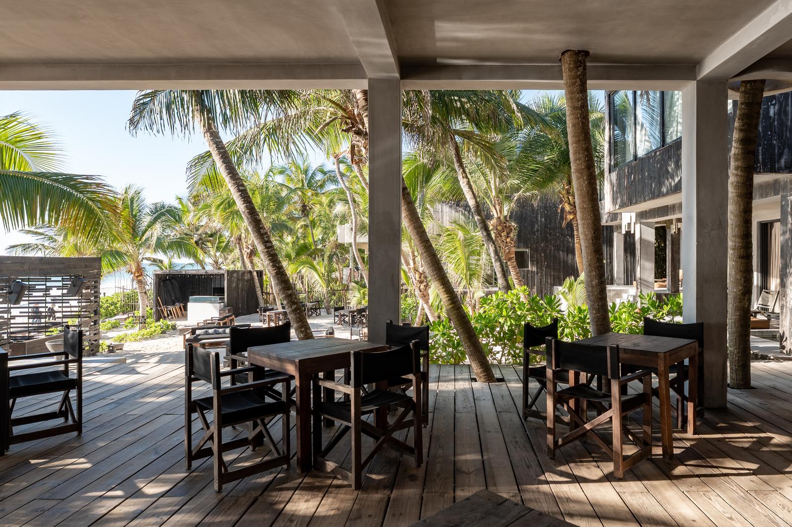 Open-air beach club dining area with wooden tables, palm trees, and ocean views in bright daylight.