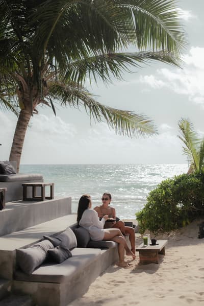 Two women relax on beach club loungers beneath palm trees beside the sparkling ocean.