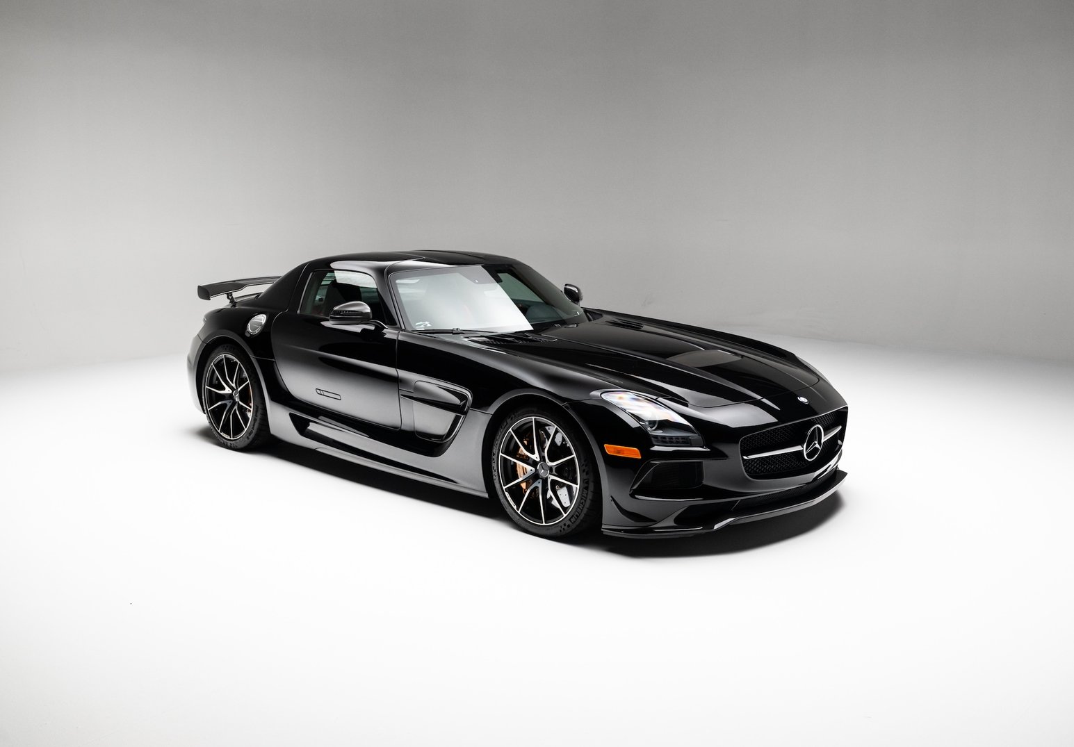 A black Mercedes-Benz SLS AMG coupe is shown in a studio on a seamless light gray background. The car is viewed from the front-left three-quarter angle with a rear wing, long hood, chrome grille emblem, and multi-spoke wheels visible.