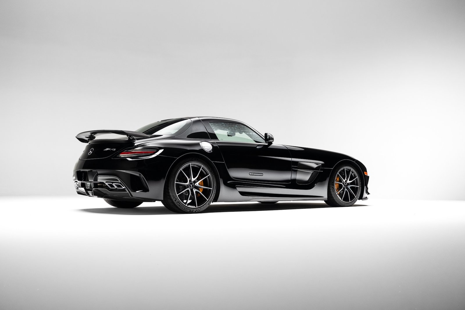 A black Mercedes-Benz SLS AMG coupe is shown in a studio on a white-to-gray seamless background. The car is viewed from the left rear three-quarter angle with long hood, coupe roofline, multi-spoke wheels, and rear taillights visible.