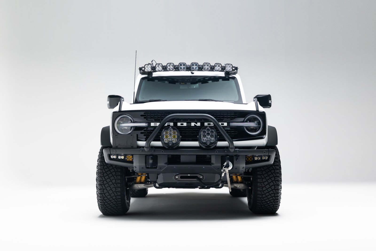 A white Ford Bronco SUV is photographed head-on in a plain studio setting. It has a black front bumper with a winch hook, auxiliary lights on the bumper and roof rack, wide off-road tires, and visible BRONCO grille lettering.