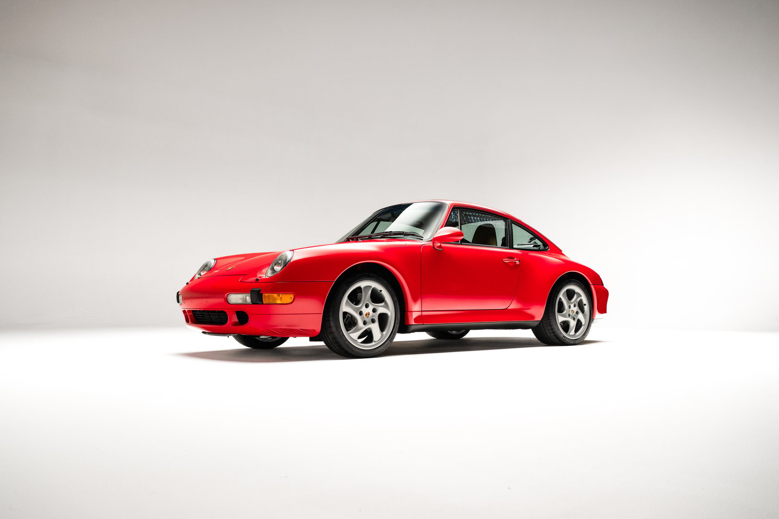 The image features a 1998 Porsche 911 Carrera S coupe in Guards Red, showcasing its sleek design and distinctive curves. The car is positioned at a front-3-4 angle, highlighting its headlights and sporty profile against 