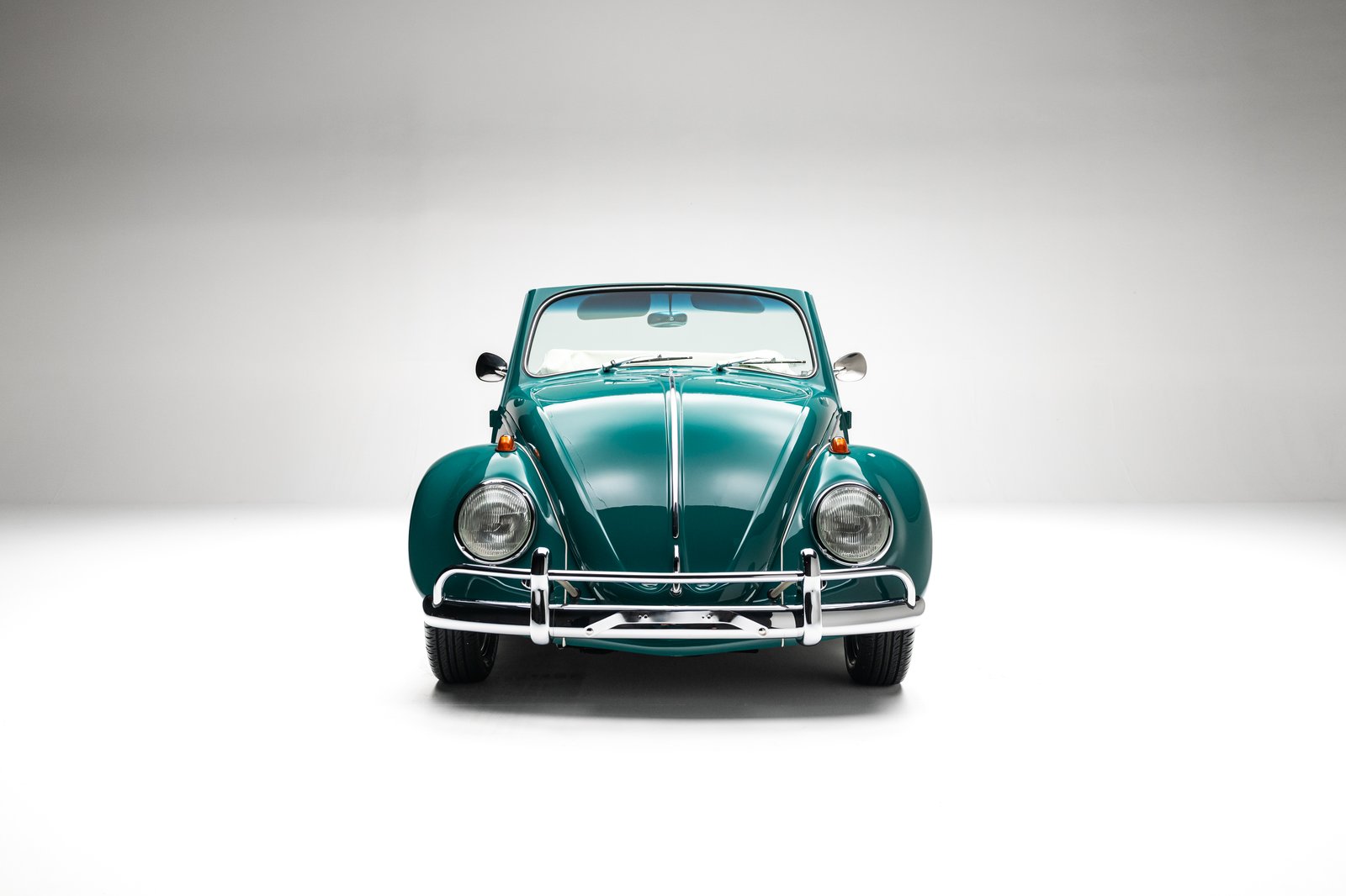 A teal 1964 Volkswagen Beetle is shown from the front in a clean white studio setting. The image highlights its round headlights, chrome bumper, hood trim, windshield, and fenders.