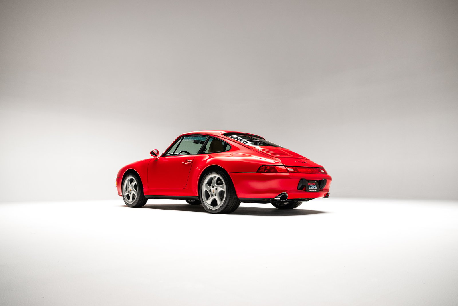 The image features a 1998 Porsche 911 Carrera S coupe in Guards Red, showcasing its sleek design and distinctive rear profile. The car is positioned in a well-lit studio setting, highlighting its polished exterior and al