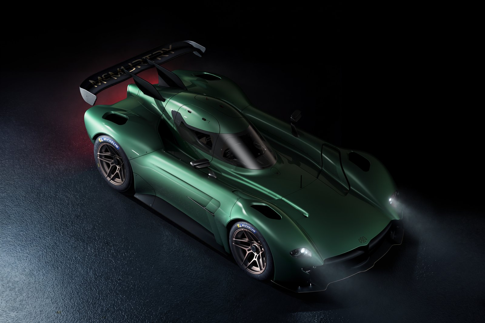 A green track-focused sports car is shown in a dark studio setting from an elevated front three-quarter view. It has illuminated front lights, exposed aerodynamic bodywork, Michelin tires, and a large rear wing with visible lettering.