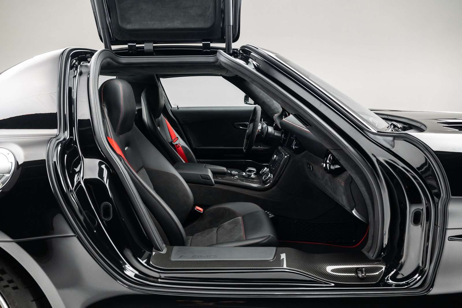 The image shows a black Mercedes-Benz SLS AMG coupe in a studio setting with the gullwing door open. The view focuses on the cabin and open side doorway, showing black seats with red seat belts and glossy exterior bodywork.