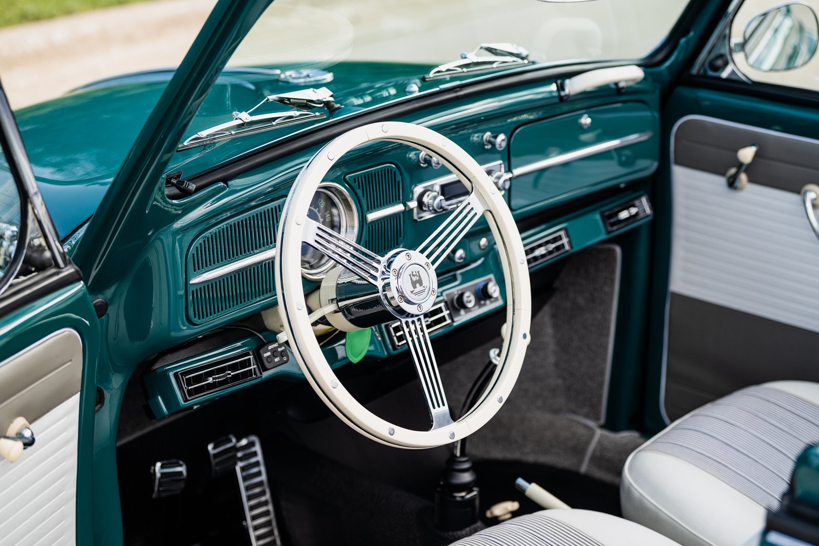 Interior view of a teal 1964 Volkswagen Beetle showing the dashboard, white steering wheel, pedals, shifter, and two-tone door panels. The photo is taken from the open side door with close framing on the driver area and 