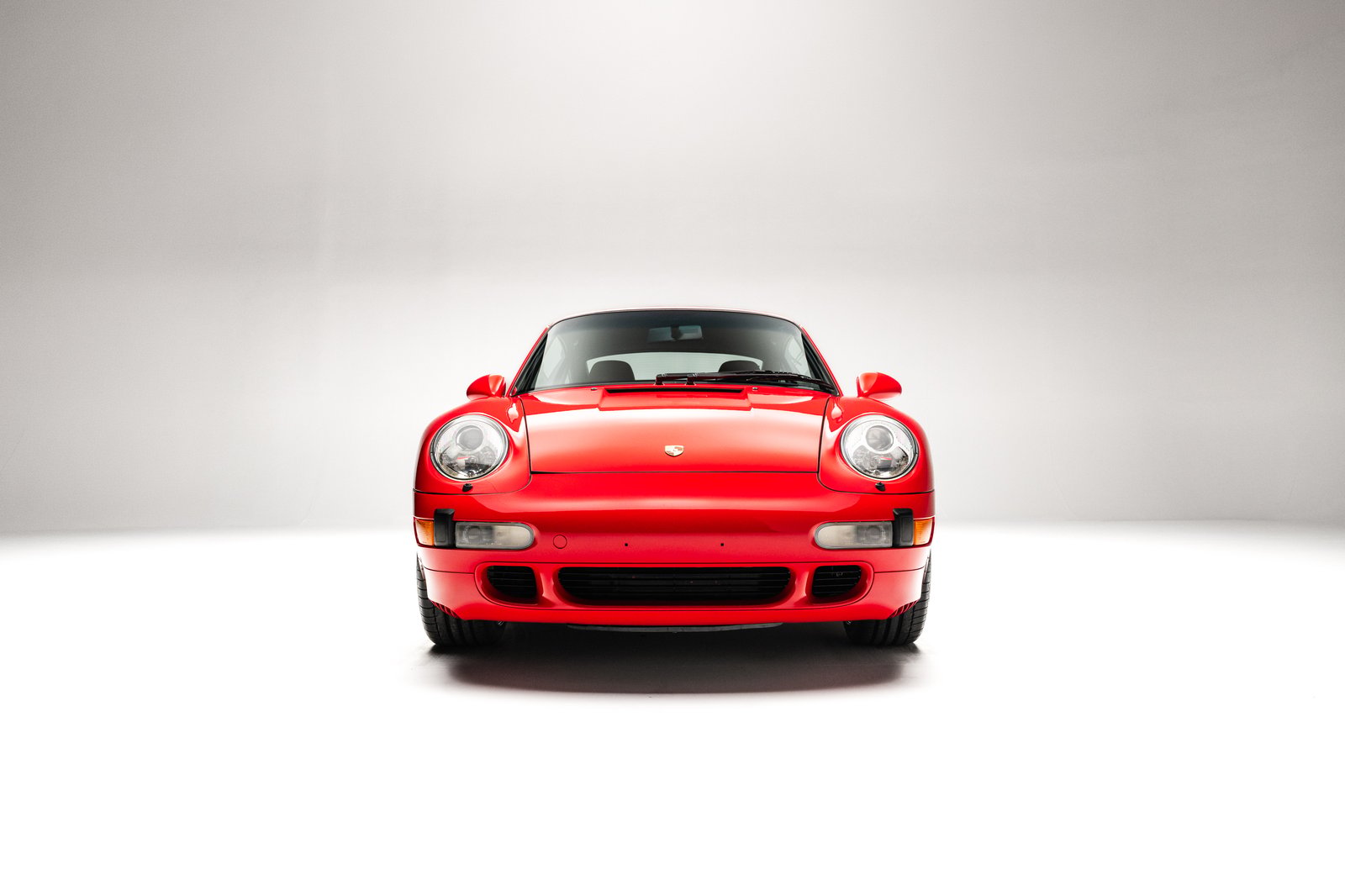 The image features a 1998 Porsche 911 Carrera S coupe in Guards Red, showcasing its front view with a clean, minimalistic background. Notable details include the round headlights and the iconic Porsche crest on the hood.