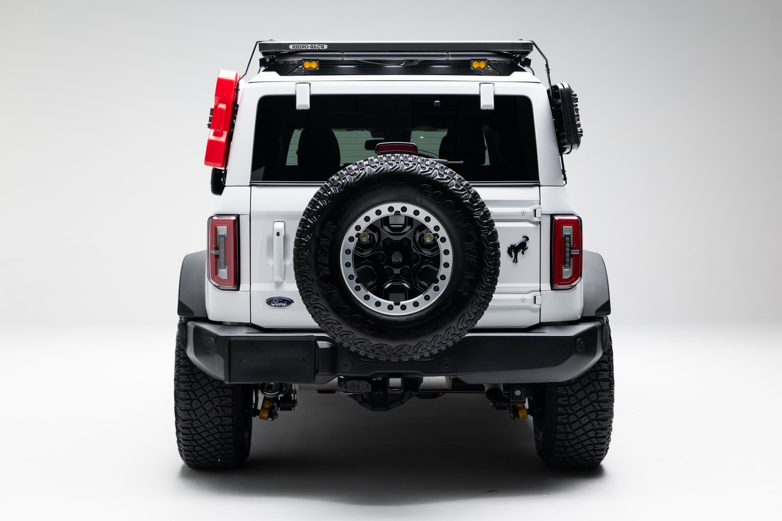 A white Ford Bronco four-door SUV is shown from the rear in a studio setting on a plain light background. The rear-mounted spare tire, roof rack with lighting, side-mounted gear, black bumper, and vertical taillights are visible.