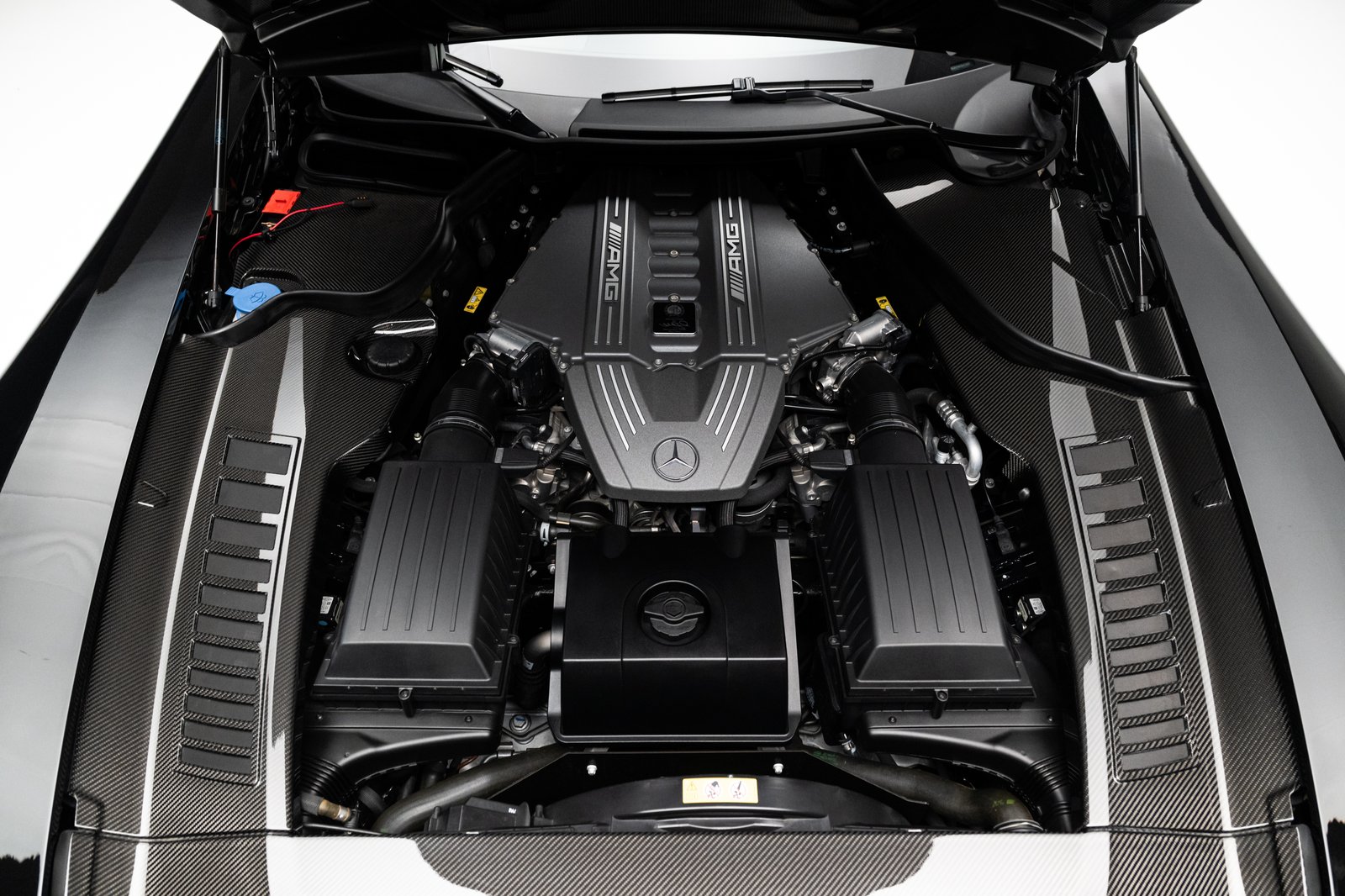 The image shows the engine bay of a black Mercedes-Benz SLS AMG with the hood open in a studio-like setting. Carbon-fiber panels, AMG-branded engine covers, intake components, and surrounding bay structure are visible in a close, overhead-oriented view.