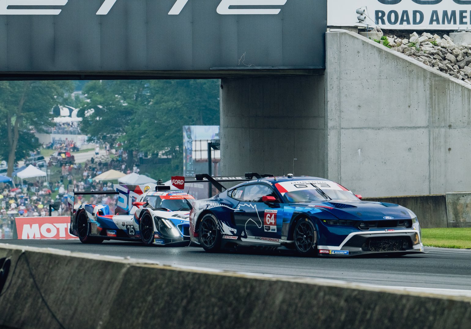 Two race cars in motion during a race at Road America, viewed from a front 3/4 angle.