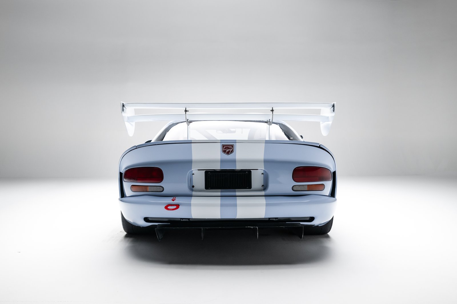 A blue Dodge Viper GTS race car with white center stripes is shown from the rear in a white studio setting. The rear view highlights oval taillights, a large adjustable wing, tow hook, rear diffuser, and wide slick tires